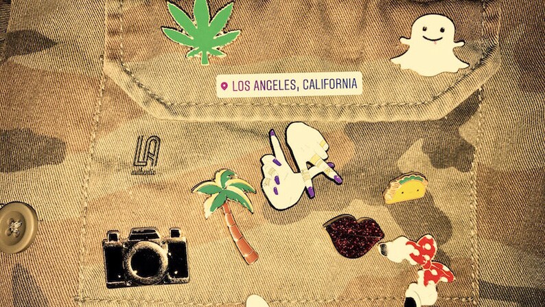 May include: A brown camouflage jacket with various pins, including a palm tree, a camera, a ghost, a taco, a red bow, a glitter lip, and a pin that says "LA" with a location pin and the text "Los Angeles, California".
