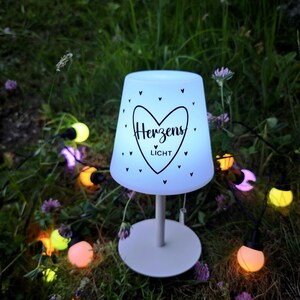 May include: A white table lamp with a conical shade featuring a black heart graphic and the text "Herzens LICHT" surrounded by small heart icons. The lamp has a thin stem and a round base, illuminated from within, set among colourful string lights.