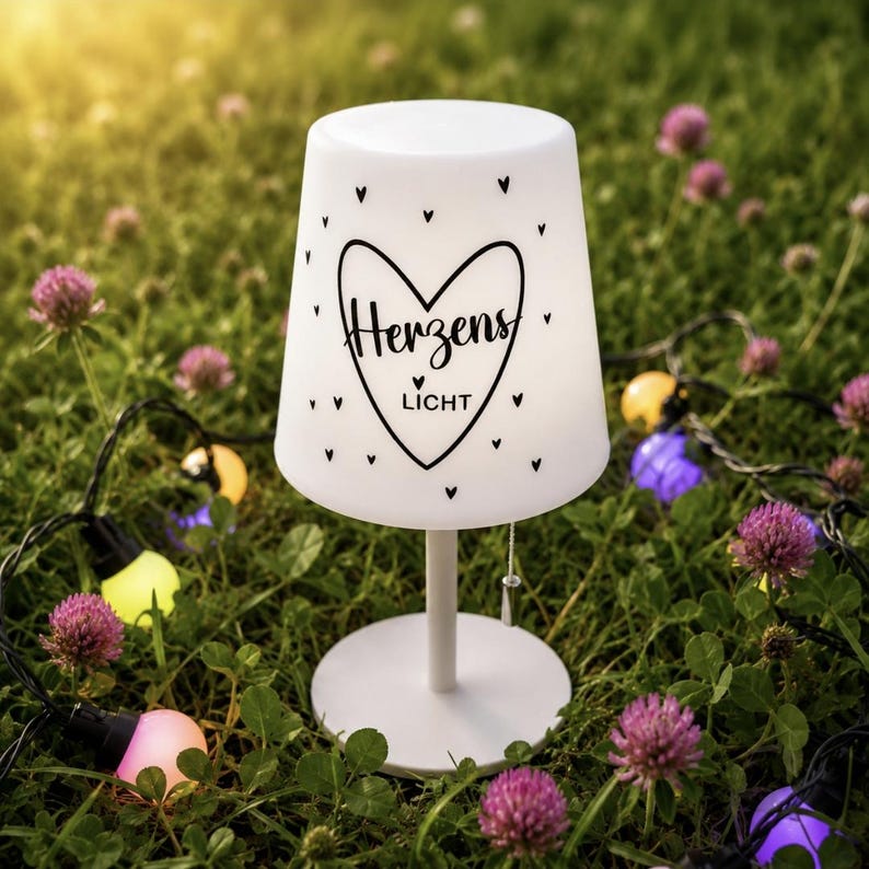 May include: A white table lamp with a tapered shade featuring a black heart graphic and the text "Herzens LICHT" surrounded by small heart icons. The lamp has a thin white stem, a round base, and a pull-chain switch hanging from the shade.