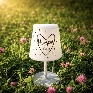 May include: A white table lamp with a conical shade featuring a black heart outline, small scattered hearts, and the text "Herzens LICHT" printed in the centre. The lamp has a thin vertical stand, a round base, and a pull chain switch.