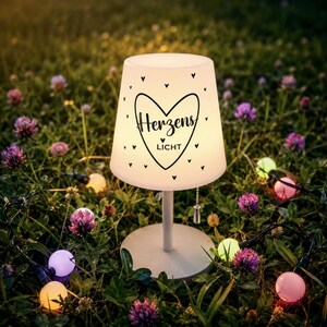 May include: A white table lamp with a conical shade featuring a black heart graphic and the text "Herzens LICHT" surrounded by small heart icons. The lamp has a thin stem, a round base, and a pull-chain switch hanging from the shade.