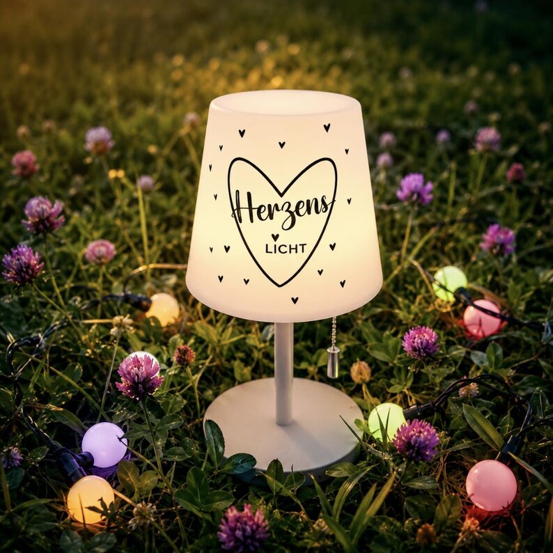 May include: A white table lamp with a conical shade featuring a black heart graphic and the text "Herzens LICHT" surrounded by small heart icons. The lamp has a thin stem, a round base, and a pull-chain switch hanging from the shade.