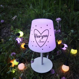 May include: A portable table lamp with a glowing purple shade featuring a black heart graphic and the text Herzens Licht. The lamp has a white base and a slender stem, surrounded by a string of small, multi-coloured spherical LED lights.