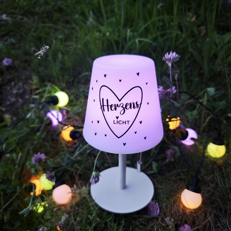 May include: A portable table lamp with a glowing purple shade featuring a black heart graphic and the text Herzens Licht. The lamp has a white base and a slender stem, surrounded by a string of small, multi-coloured spherical LED lights.