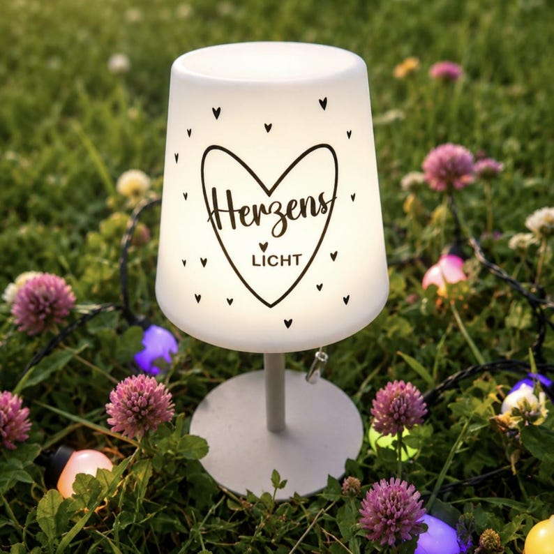 May include: A white, tapered, illuminated table lamp with a round base. The lamp shade features a black graphic of a heart containing the text "Herzens LICHT" and small scattered heart icons. A pull cord switch hangs from the side of the shade.