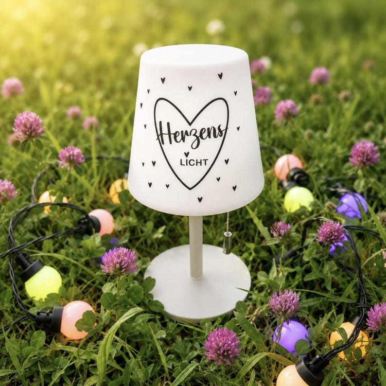 May include: A white table lamp with a tapered shade featuring a black heart graphic and the text "Herzens LICHT" surrounded by small heart icons. The lamp has a simple cylindrical stem and a round base, standing upright on a flat surface.