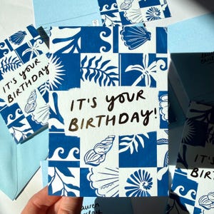 K&ouml;nnte beinhalten: Eine rechteckige Gru&szlig;karte mit einem blau-wei&szlig;en Kachelmuster aus Muscheln, Korallen und Wellenmotiven. In der Mitte der Karte befindet sich ein wei&szlig;es Rechteck mit dem Text IT'S YOUR BIRTHDAY! in einer dunklen, handschriftlichen Schriftart.