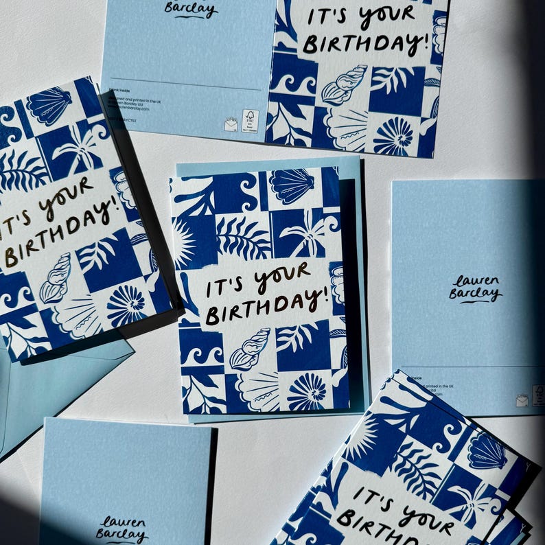 K&ouml;nnte beinhalten: Eine Sammlung rechteckiger Gru&szlig;karten mit einem blau-wei&szlig;en Kachelmuster und Illustrationen von Muscheln und Palmbl&auml;ttern. Jede Karte zeigt den handgeschriebenen schwarzen Text IT'S YOUR BIRTHDAY! sowie den Namen Lauren Barclay.