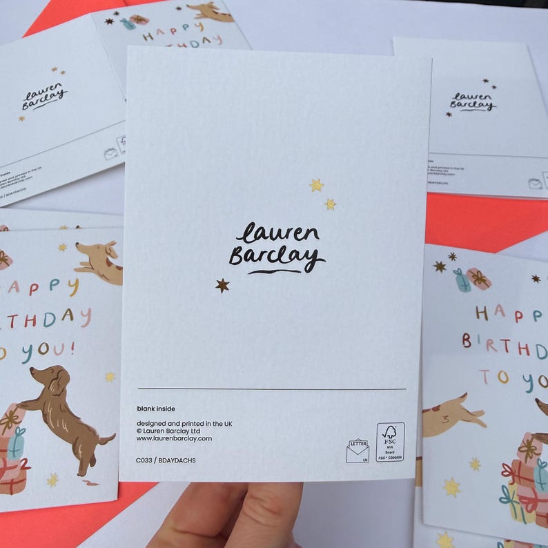 Puede incluir: Una tarjeta de felicitaci&oacute;n blanca con el texto negro 'lauren Barclay' centrado y peque&ntilde;as estrellas doradas. El reverso incluye el texto 'blank inside', 'designed and printed in the UK', 'laurenbarclay.com', 'C033 / BDAYDACHS' y un logotipo FSC.