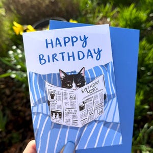 May include: A white birthday greeting card featuring a black and white cat reading a newspaper titled Birthday Mews while lying on blue and white striped bedding. The text Happy Birthday is printed in blue at the top of the card.
