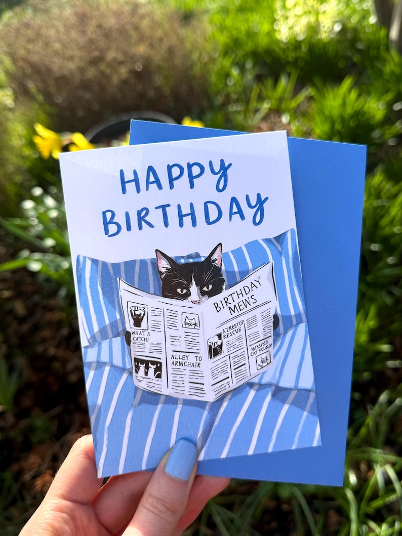 May include: A white birthday greeting card featuring a black and white cat reading a newspaper titled Birthday Mews while lying on blue and white striped bedding. The text Happy Birthday is printed in blue at the top of the card.