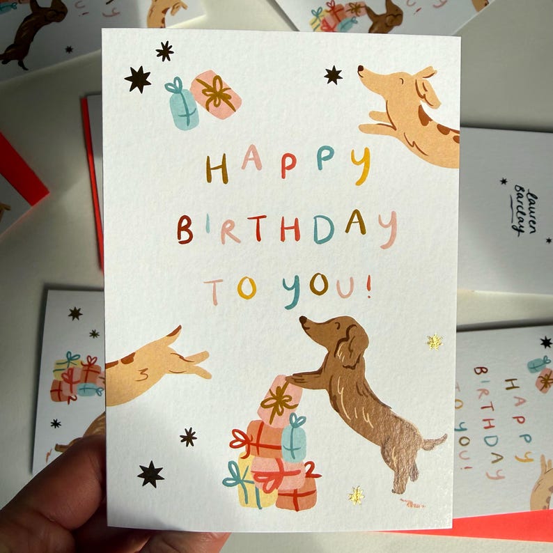 Puede incluir: Una tarjeta de felicitaci&oacute;n blanca con el texto HAPPY BIRTHDAY TO YOU en letras multicolores. El dise&ntilde;o incluye dos perros salchicha marrones, uno saltando y otro de pie junto a una pila de cajas de regalo coloridas, con peque&ntilde;as estrellas negras.