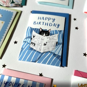 May include: A greeting card featuring a black and white cat reading a newspaper titled Birthday Mews while lying in a blue and white striped bed. The text Happy Birthday is printed in blue above the cat. The card is set against a white background.