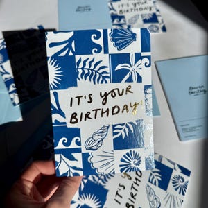 K&ouml;nnte beinhalten: Eine rechteckige Gru&szlig;karte mit einem blau-wei&szlig;en Kachelmuster aus Muscheln und Bl&auml;ttern. In der Mitte der Karte befindet sich der goldene Schriftzug IT'S YOUR BIRTHDAY! auf einem wei&szlig;en rechteckigen Hintergrund, umgeben von dem blauen Muster.