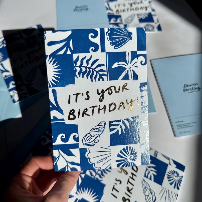 K&ouml;nnte beinhalten: Eine rechteckige Gru&szlig;karte mit einem blau-wei&szlig;en Kachelmuster aus Muscheln und Bl&auml;ttern. In der Mitte der Karte befindet sich der goldene Schriftzug IT'S YOUR BIRTHDAY! auf einem wei&szlig;en rechteckigen Hintergrund, umgeben von dem blauen Muster.