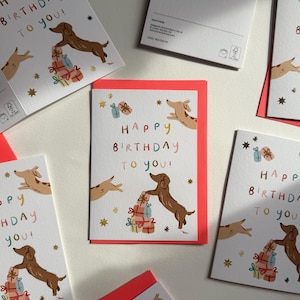 Puede incluir: Una tarjeta de felicitaci&oacute;n blanca con la ilustraci&oacute;n de un perro salchicha marr&oacute;n, una pila de regalos coloridos y el texto "Happy Birthday to you!" en letras multicolores. Estrellas doradas decoran la tarjeta, que incluye un sobre rojo.