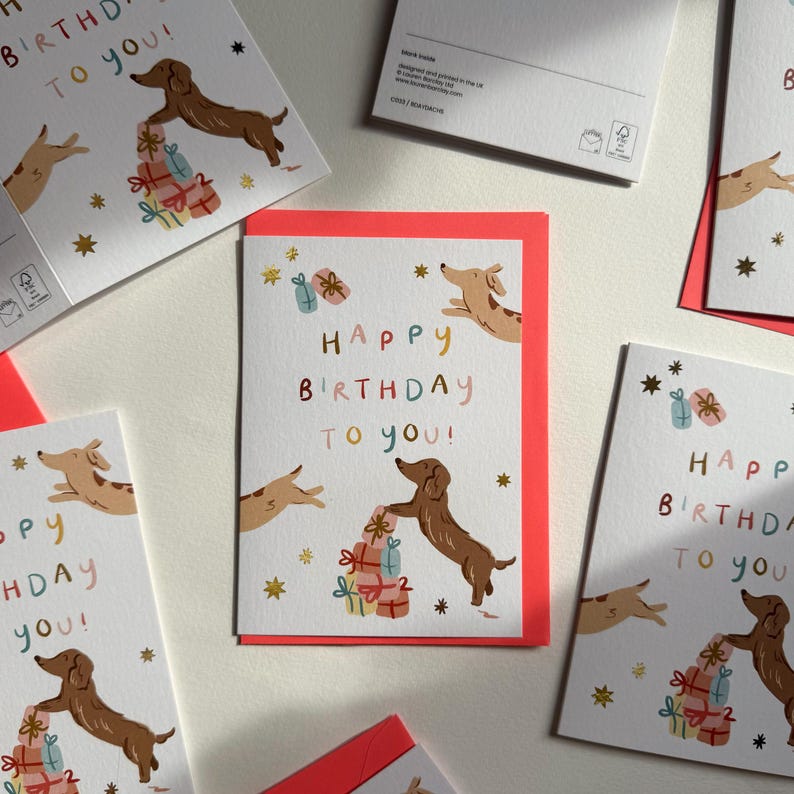Puede incluir: Una tarjeta de felicitaci&oacute;n blanca con la ilustraci&oacute;n de un perro salchicha marr&oacute;n, una pila de regalos coloridos y el texto "Happy Birthday to you!" en letras multicolores. Estrellas doradas decoran la tarjeta, que incluye un sobre rojo.