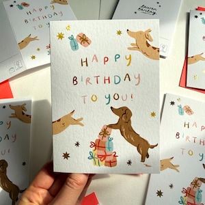Puede incluir: Una tarjeta de felicitaci&oacute;n de cumplea&ntilde;os blanca con perros salchicha marrones, cajas de regalo coloridas y estrellas doradas. El texto Happy Birthday to you aparece en letras multicolores. La tarjeta es de papel texturizado.