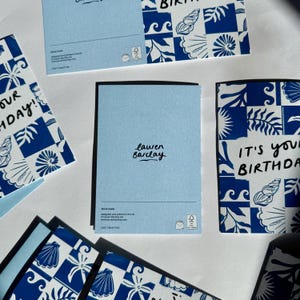 K&ouml;nnte beinhalten: Eine Sammlung von Gru&szlig;karten mit einem blau-wei&szlig;en Kachelmuster aus Muscheln und Bl&auml;ttern. Die Karten zeigen den Text "IT'S YOUR BIRTHDAY!" in schwarzer Schrift. Eine Karte zeigt die R&uuml;ckseite mit dem Markennamen "lauren barclay".