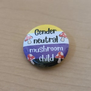 May include: A circular badge with the text "Gender neutral mushroom child". The badge has a yellow top, a purple middle, and a black bottom. It features cartoon mushrooms with red caps and white spots. The badge is a pin.