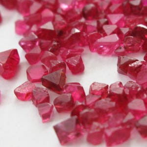 May include: A collection of loose red ruby gemstones, some with a triangular shape, scattered on a white surface.