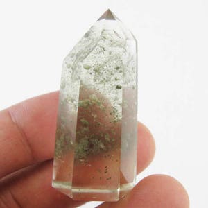 Puede incluir: Una punta de cristal de cuarzo transparente con inclusiones verdes. El cristal es puntiagudo en la parte superior y tiene una base plana. Mide aproximadamente 5 cm de largo.