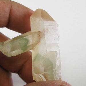Puede incluir: Un cristal de cuarzo transparente con inclusiones verdes. El cristal está doblemente terminado y tiene una superficie natural y rugosa.