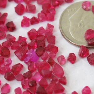 May include: A close-up of a collection of small, red, faceted gemstones. The gemstones are scattered on a white surface, with a US dime for scale.