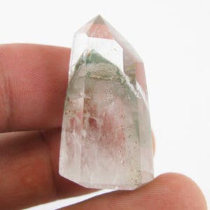 Puede incluir: Una punta de cristal de cuarzo transparente con inclusiones verdes. El cristal es puntiagudo en la parte superior y tiene una base plana. Mide aproximadamente 5 cm de largo.