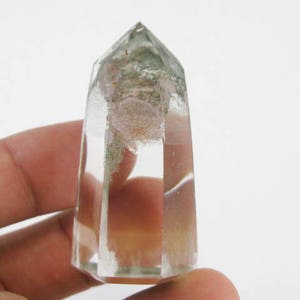 Puede incluir: Una punta de cristal de cuarzo transparente con inclusiones verdes. El cristal está facetado y tiene una punta puntiaguda. Mide aproximadamente 5 cm de alto.