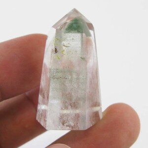 Puede incluir: Una punta de cristal de cuarzo transparente con inclusiones verdes. El cristal está facetado y tiene una punta puntiaguda. Mide aproximadamente 5 cm de alto.