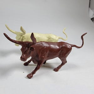 May include: Two toy longhorn bulls. The foreground bull is a dark brown color, and the background bull is a light cream color. Both have large horns and are posed in a walking stance. The toys are made of plastic.