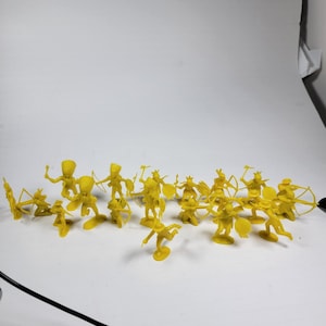 May include: A collection of yellow plastic toy soldiers in various poses, including archers, warriors with shields, and figures with raised weapons. The figures are arranged on a white surface.