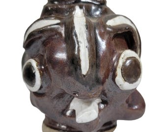 May include: A brown ceramic face jug with white accents. The jug has large, round eyes with white rims and a white stripe down the center. The jug has a small, white, rectangular shape for a mouth.