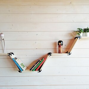 K&ouml;nnte beinhalten: Ein Holzregalsystem mit einem einzigartigen Zickzack-Design, bestehend aus drei Regalbrettern. Das obere Regal enth&auml;lt drei kleine Topfpflanzen, das mittlere Regal enth&auml;lt eine Puppe und das untere Regal enth&auml;lt B&uuml;cher.