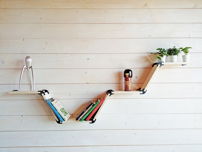 K&ouml;nnte beinhalten: Ein Holzregalsystem mit einem einzigartigen Zickzack-Design, bestehend aus drei Regalbrettern. Das obere Regal enth&auml;lt drei kleine Topfpflanzen, das mittlere Regal enth&auml;lt eine Puppe und das untere Regal enth&auml;lt B&uuml;cher.