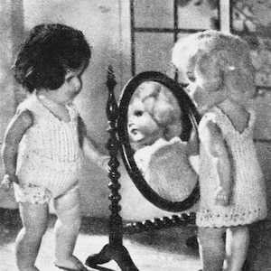 May include: A black and white photograph of a young child looking at their reflection in a vintage oval mirror. The child is wearing a white knitted dress and is standing in front of a wooden stand with a carved design.