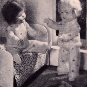 May include: Two dolls dressed in pajamas, one sitting on a bed and the other standing next to the bed. The doll sitting on the bed is wearing a white and gray striped pajama top and pants. The doll standing next to the bed is wearing a white and gray striped pajama top and pants with a white collar.
