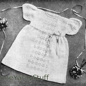 Peut inclure: Une robe de bébé en tricot blanc avec un motif de dentelle et un ruban de satin noué à la taille. La robe est posée sur une surface texturée avec deux petits bouquets de fleurs en crochet blanc.