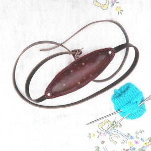 May include: A brown leather guitar strap with a padded center, featuring decorative rivets and red stitching. The strap is shown on a white surface with a blue knitted item and knitting needles.
