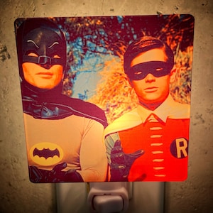May include: A nightlight featuring a vintage image of Batman and Robin. Batman is wearing a black mask and cape, while Robin is wearing a red and yellow costume with the letter "R" on his chest. The background is a blurred image of trees and foliage.