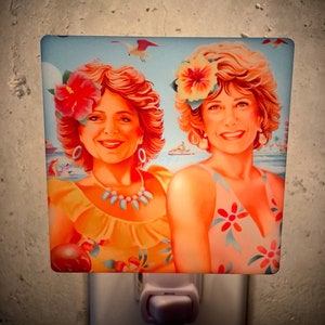 May include: Two women with bright smiles are wearing colourful summer dresses and floral headbands. The image is a nightlight with a blue background and a white switch.