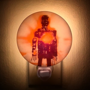 May include: A nightlight with a white base and a round, translucent orange dome. The dome features a silhouette of a person with a ladder, printed in shades of red and orange.