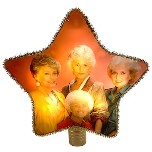 May include: A gold star-shaped Christmas tree topper featuring a picture of the four Golden Girls. The star has a gold fringe border.