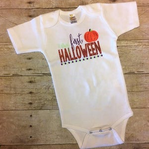 May include: White baby onesie with the text "My first Halloween" in orange glitter with a small orange pumpkin graphic above the text.