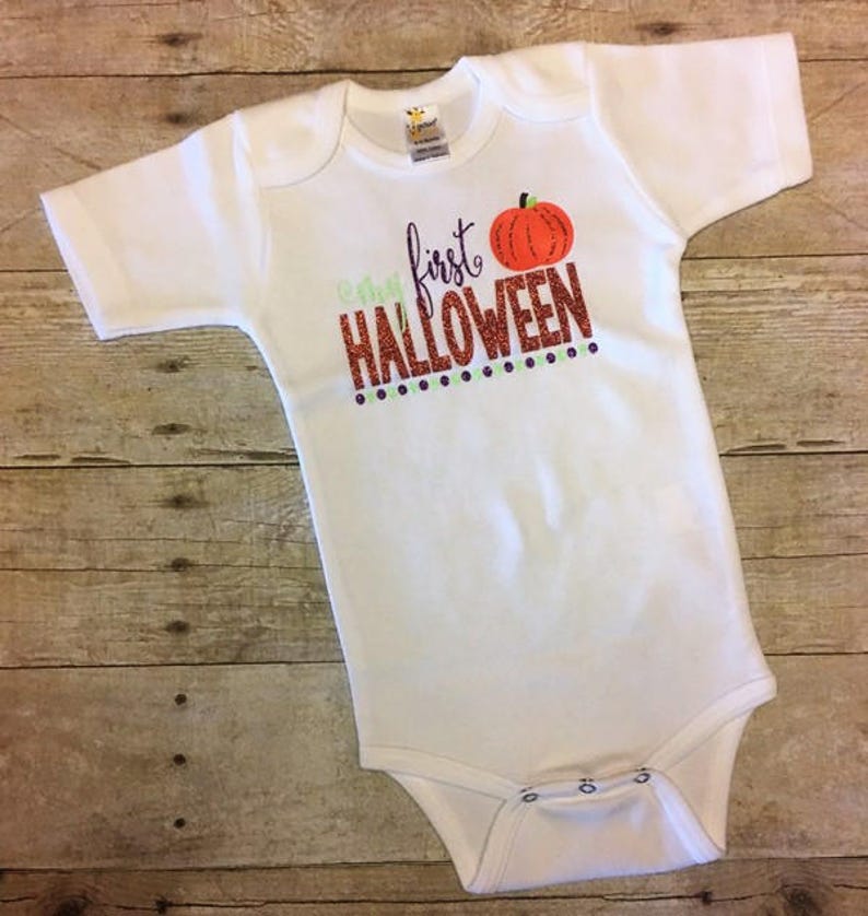 May include: White baby onesie with the text "My first Halloween" in orange glitter with a small orange pumpkin graphic above the text.