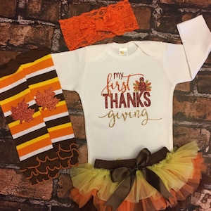 Pu&ograve; includere: Un set di abbigliamento per neonati per il Ringraziamento. Include un body bianco a maniche lunghe con il testo "my first THANKS giving" in glitter dorato e rosso, un gambaletto a righe marroni e arancioni con foglie glitterate, una gonna di tulle marrone con strati arancioni, gialli e marroni, e una fascia per capelli in pizzo arancione con un fiocco.