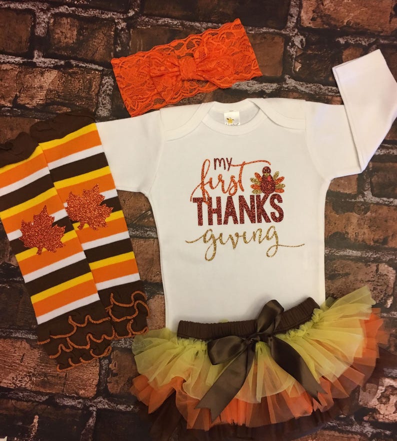 Pu&ograve; includere: Un set di abbigliamento per neonati per il Ringraziamento. Include un body bianco a maniche lunghe con il testo "my first THANKS giving" in glitter dorato e rosso, un gambaletto a righe marroni e arancioni con foglie glitterate, una gonna di tulle marrone con strati arancioni, gialli e marroni, e una fascia per capelli in pizzo arancione con un fiocco.
