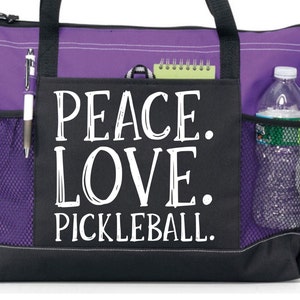 May include: Purple and black tote bag with a mesh pocket and a zippered pocket. The bag has a white graphic that says "Peace. Love. Pickleball."