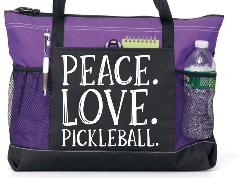 May include: Purple and black tote bag with a mesh pocket and a zippered pocket. The bag has a white graphic that says "Peace. Love. Pickleball."